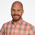 A headshot of AJ Bruno wearing a plaid gray and coral colored shirt.
